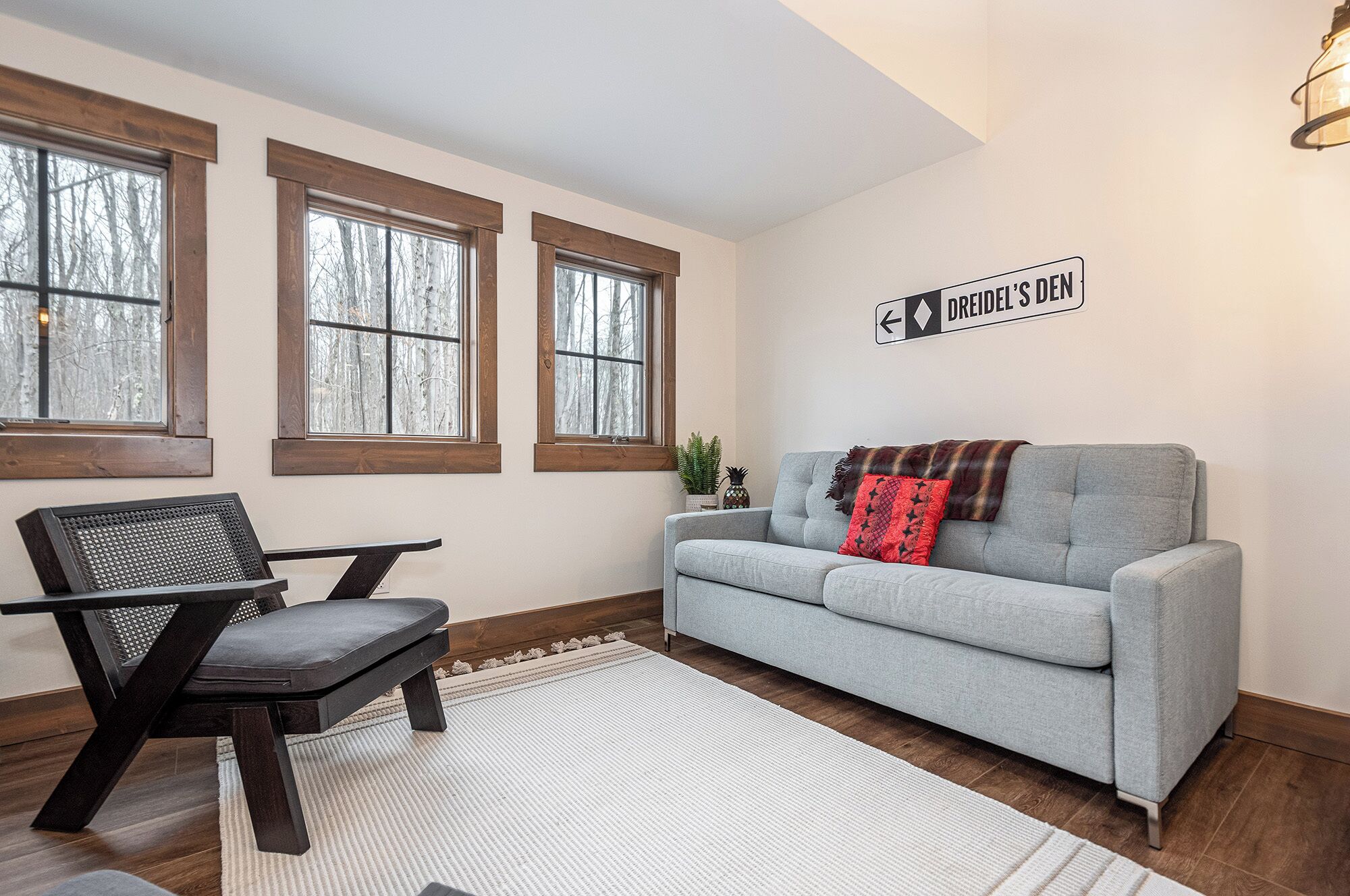 A living room with a gray sofa, a wooden chair, and a rug. There are three windows with wooden frames, and a sign on the wall.