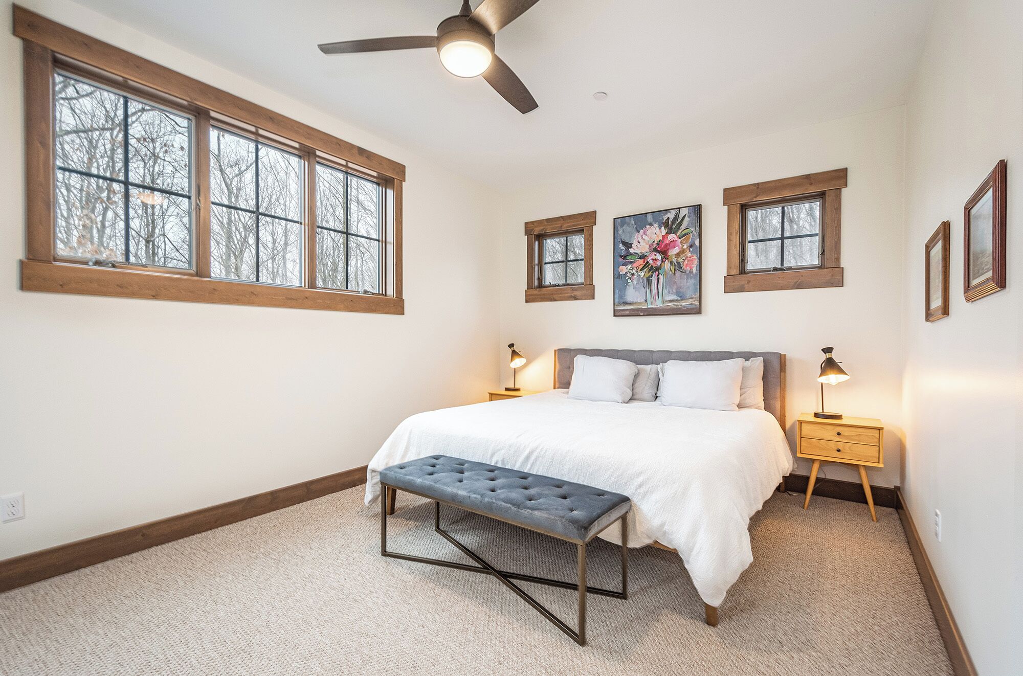 A bedroom with a bed, two bedside tables, a bench, and three windows.