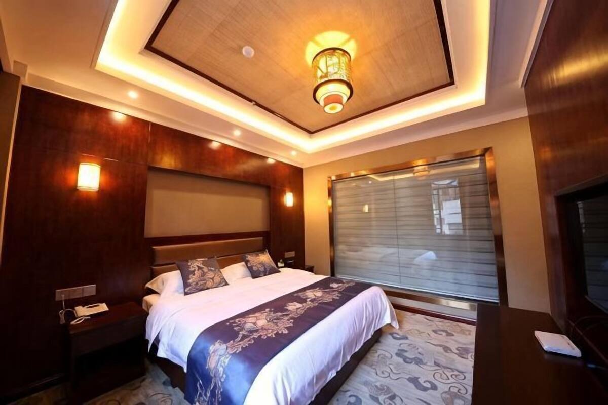 A modern hotel room with a large bed, bedside tables, a TV, and a large window with blinds.