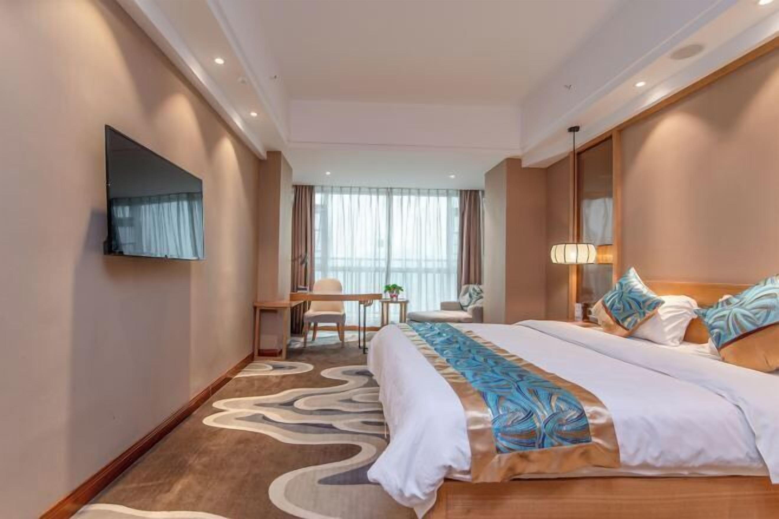 A hotel room with a large bed, a desk, a chair, a TV mounted on the wall, and a window with curtains.