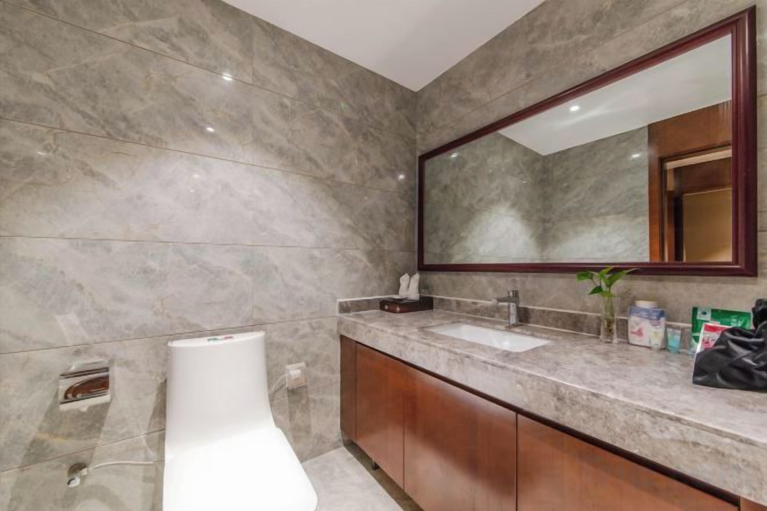 Un baño moderno con pared de mármol, un espejo grande, un lavamanos blanco y armarios de madera.