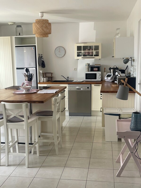 Una cocina moderna con armarios blancos, encimera de madera, una mesa de comedor con sillas, microondas, lavavajillas y una lámpara colgante.