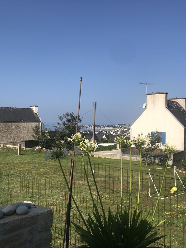 Un jardín con un muro de piedra, una valla metálica y vistas a casas y agua en la distancia.