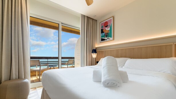 A hotel room with a large bed, a view of the beach, and a balcony with chairs.
