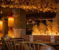 A restaurant interior with a long table set for dining, a bar area with shelves of bottles, and a decorative ceiling with foliage.
