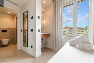 Baño de hotel con ducha, inodoro, lavamanos y un espejo grande. Hay una ventana con vistas a edificios.