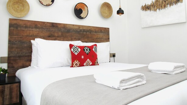 A neatly made bed with white linens and a red patterned pillow, a wooden headboard, and a wall decorated with woven wall art and pendant lights. - Zendo Suite (Nairobi)
