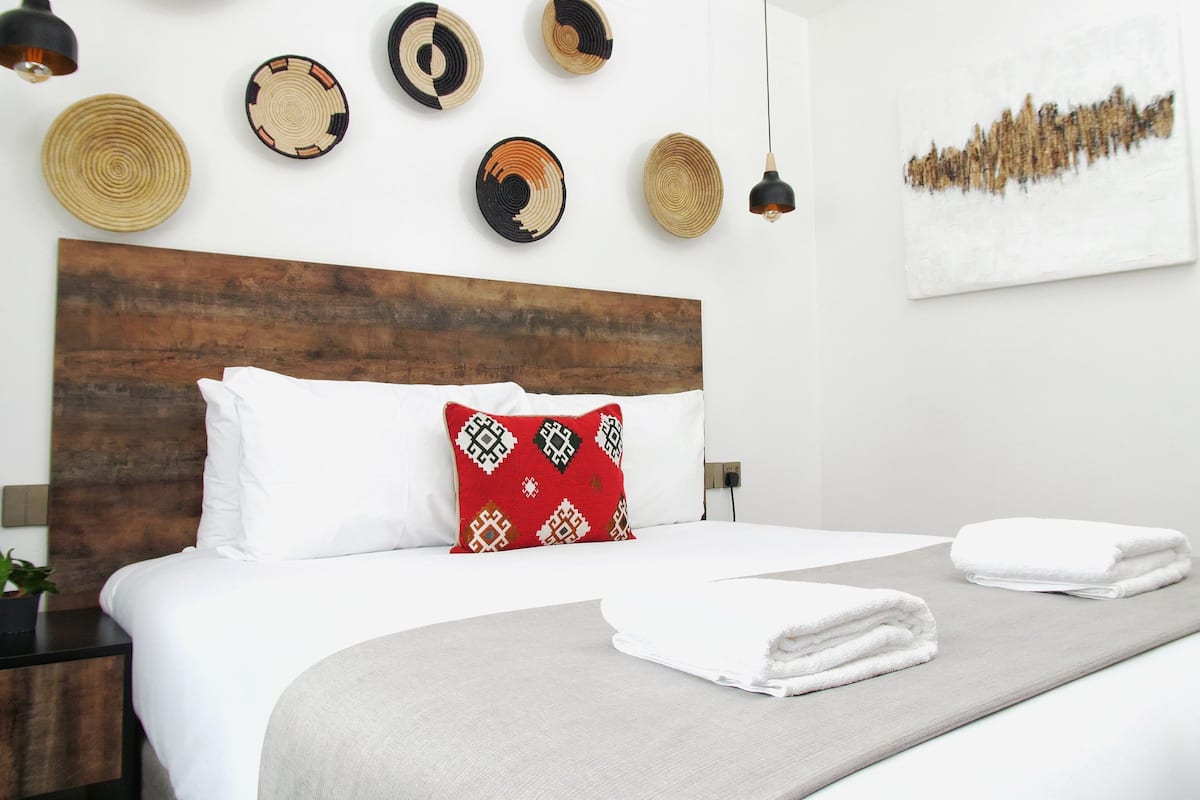 A neatly made bed with white linens and a red patterned pillow, a wooden headboard, and a wall decorated with woven wall art and pendant lights.