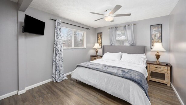 Una camera da letto con un letto, un ventilatore a soffitto, due comodini con lampade, una TV a parete e una finestra con tende.