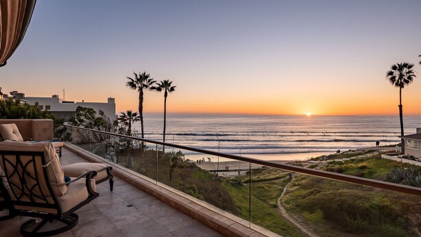 Un balcon offrant une vue sur le coucher de soleil sur l’océan, avec des palmiers et une chaise.