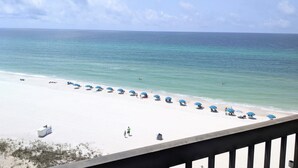 A beach with white sand and clear blue water. There are blue umbrellas and a few people on the beach.