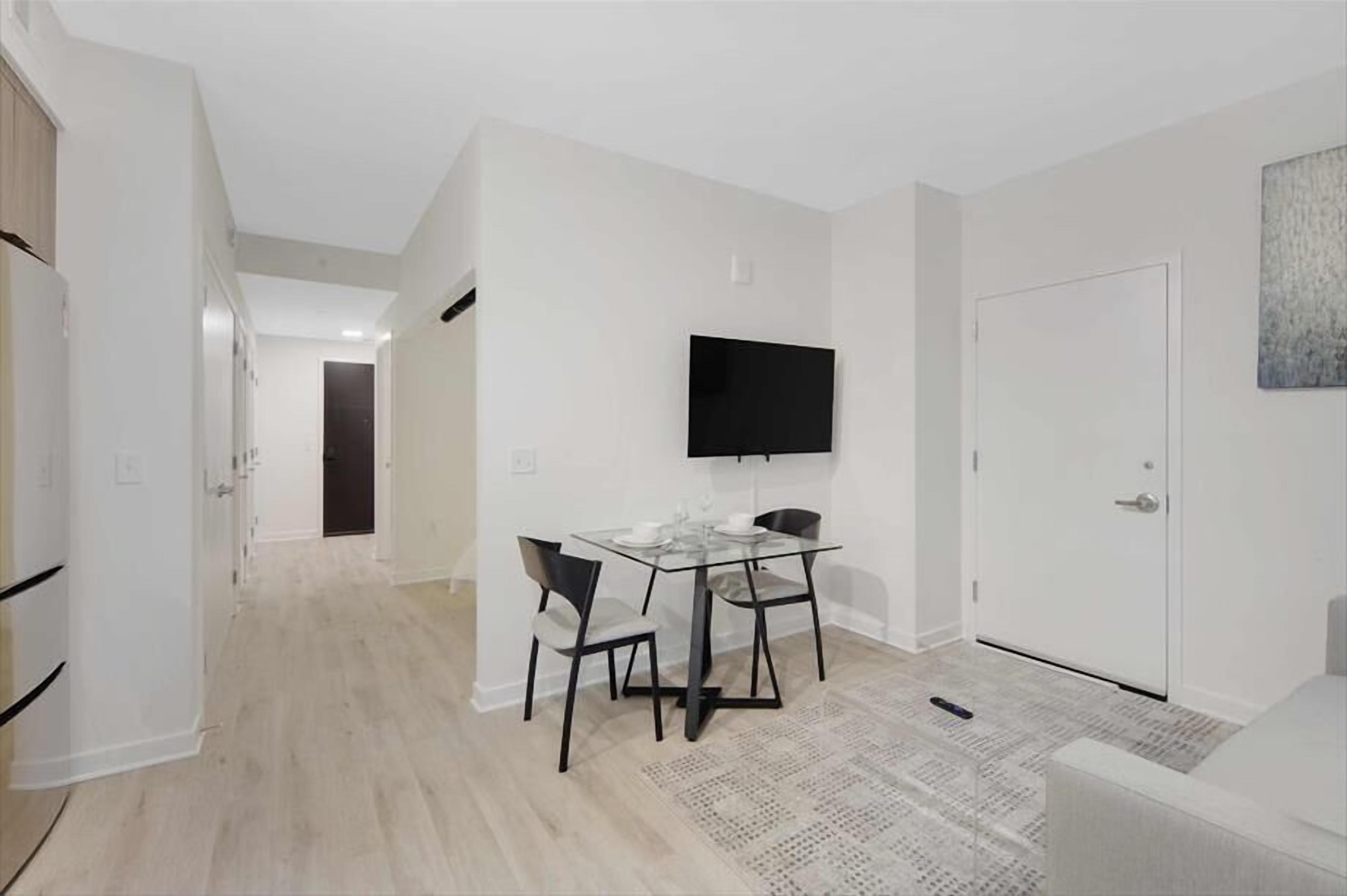 Un salon moderne et minimaliste, avec une table à manger en verre, deux chaises, un téléviseur à écran plat fixé au mur et une porte blanche.
