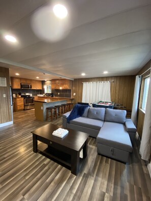 Een moderne woonkamer met een hoekbank, een salontafel en een keukenhoek met houten kasten en een magnetron.