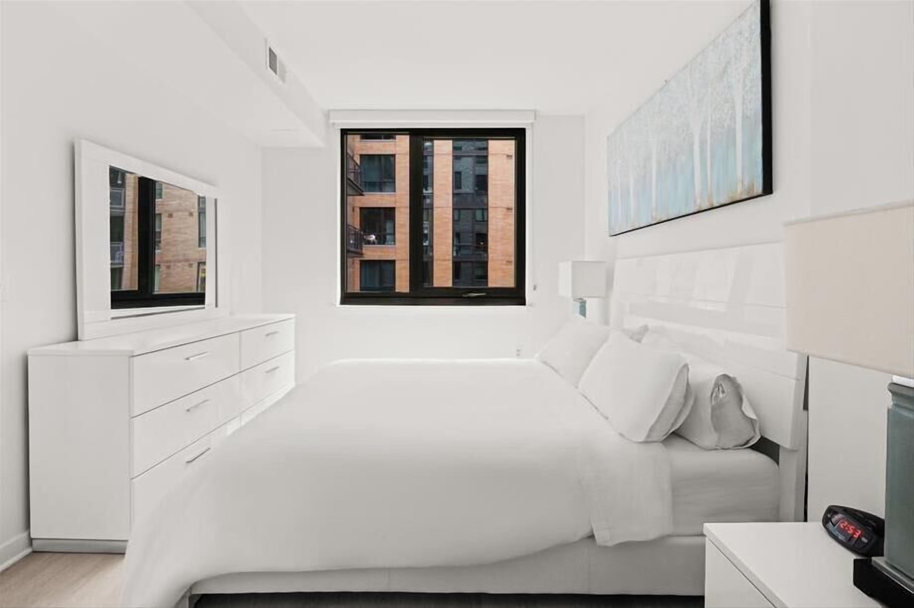 Un dormitorio moderno con una cama grande, un aparador, una mesita de noche, un televisor montado en la pared y una ventana con vistas a edificios.