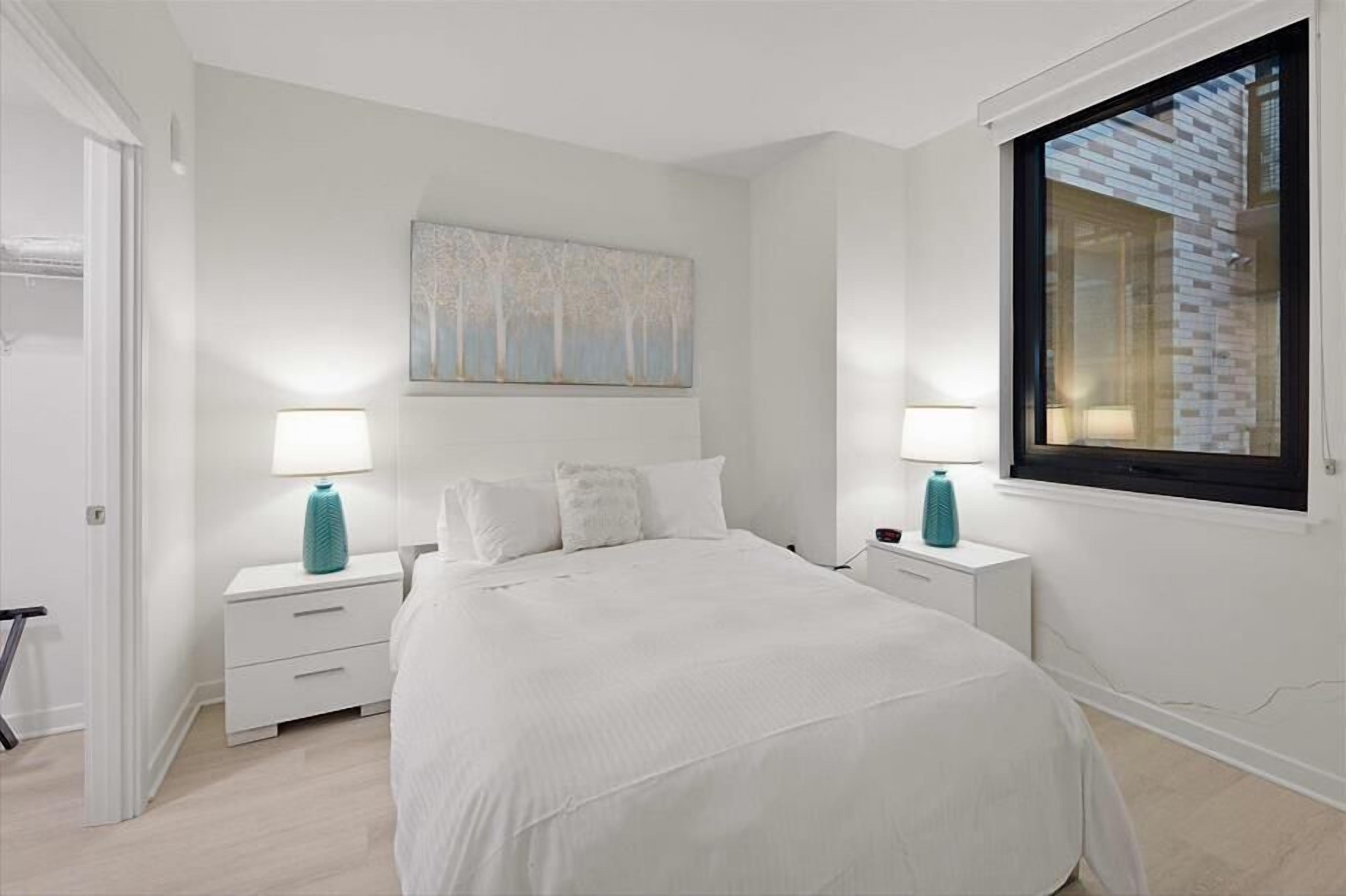 A bedroom with a large bed, two bedside tables with lamps, a window with a view of a brick building, and a painting on the wall.