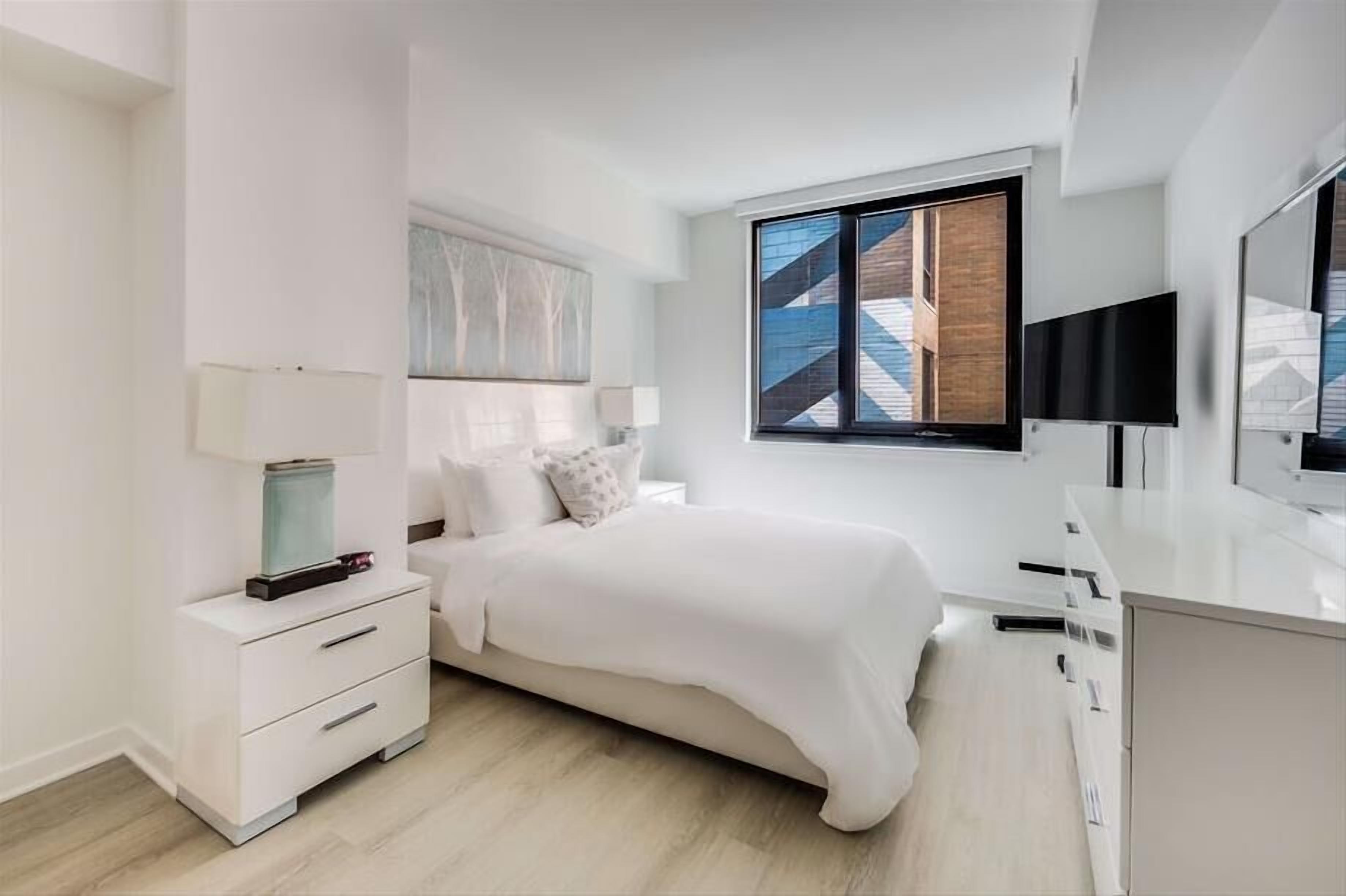 A modern bedroom with a bed, a nightstand, a TV, and a window with a view of a building.