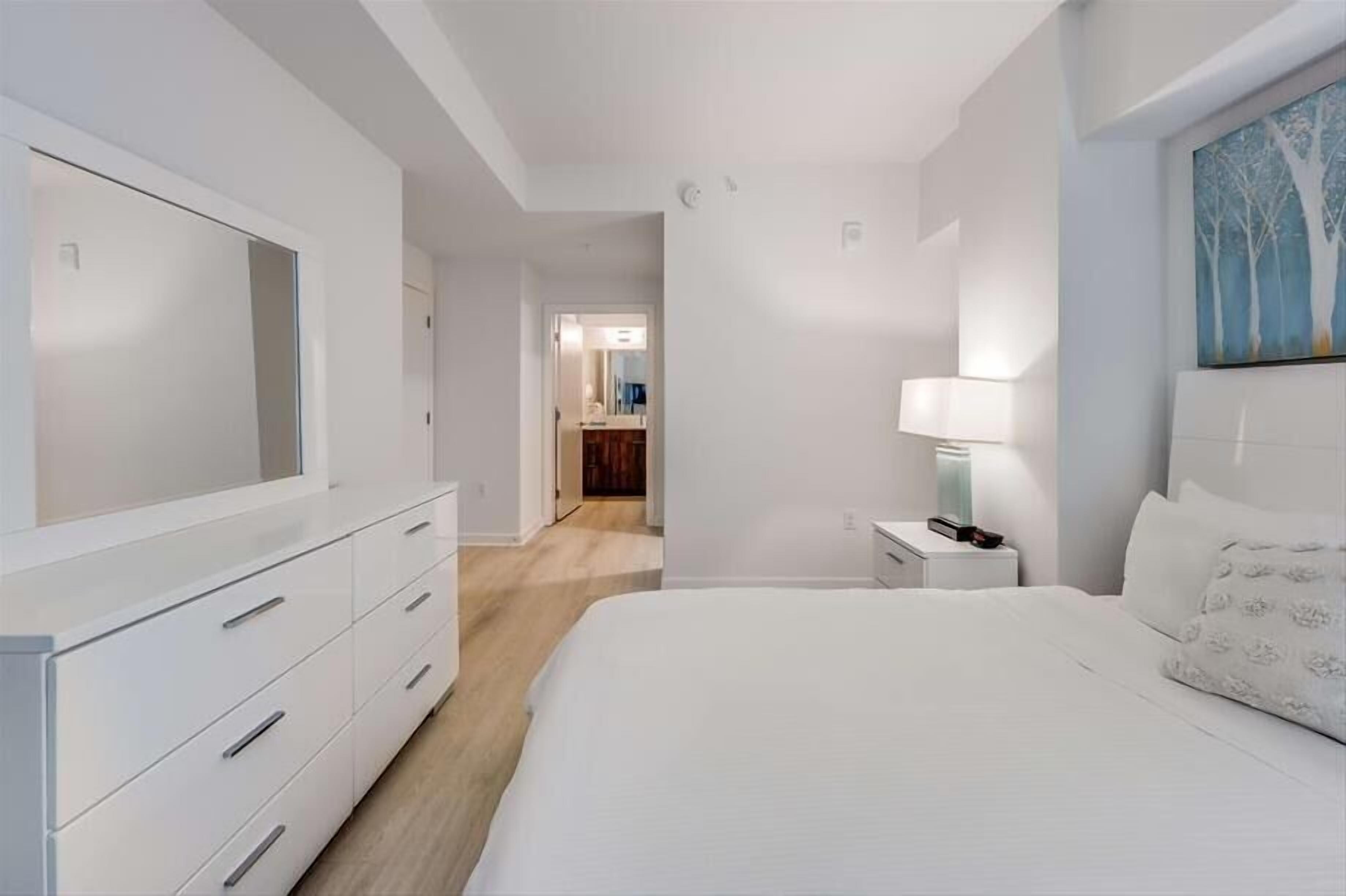 A modern hotel room with a large bed, a dresser, a nightstand, and a painting on the wall.