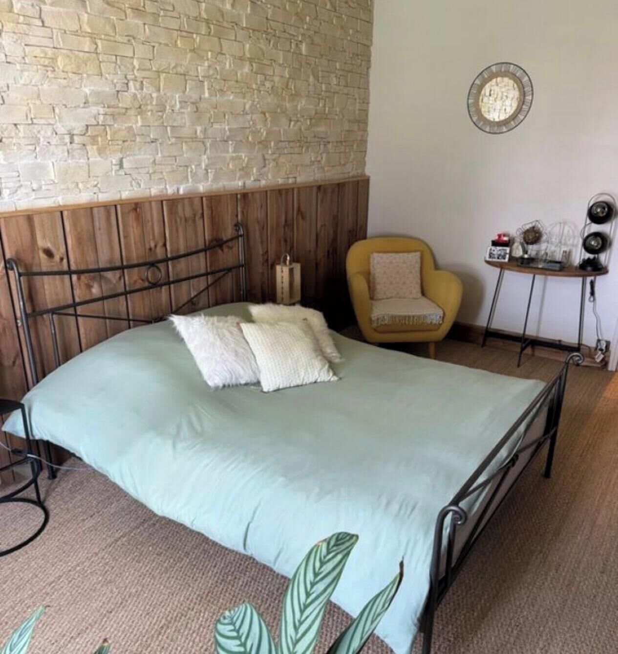 A room with a bed, a wooden headboard, a yellow armchair, a small table with a lamp, and a wall clock.