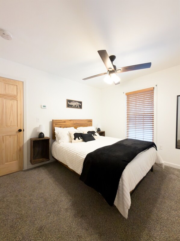 A bedroom with a bed, a ceiling fan, a nightstand, a wooden door, a window with blinds, and a wall-mounted light fixture. - Family Retreat | Game Room + Firepit | Sleeps 10 (Conway)