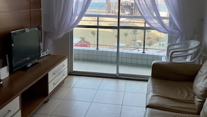 A living room with a TV on a wooden stand, a beige sofa, and a large window with sheer curtains.