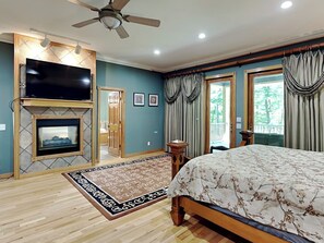 Ein Schlafzimmer mit einem großen Bett, einem Fernseher über einem Kamin, einem Deckenventilator und Blick ins Freie durch ein Fenster.