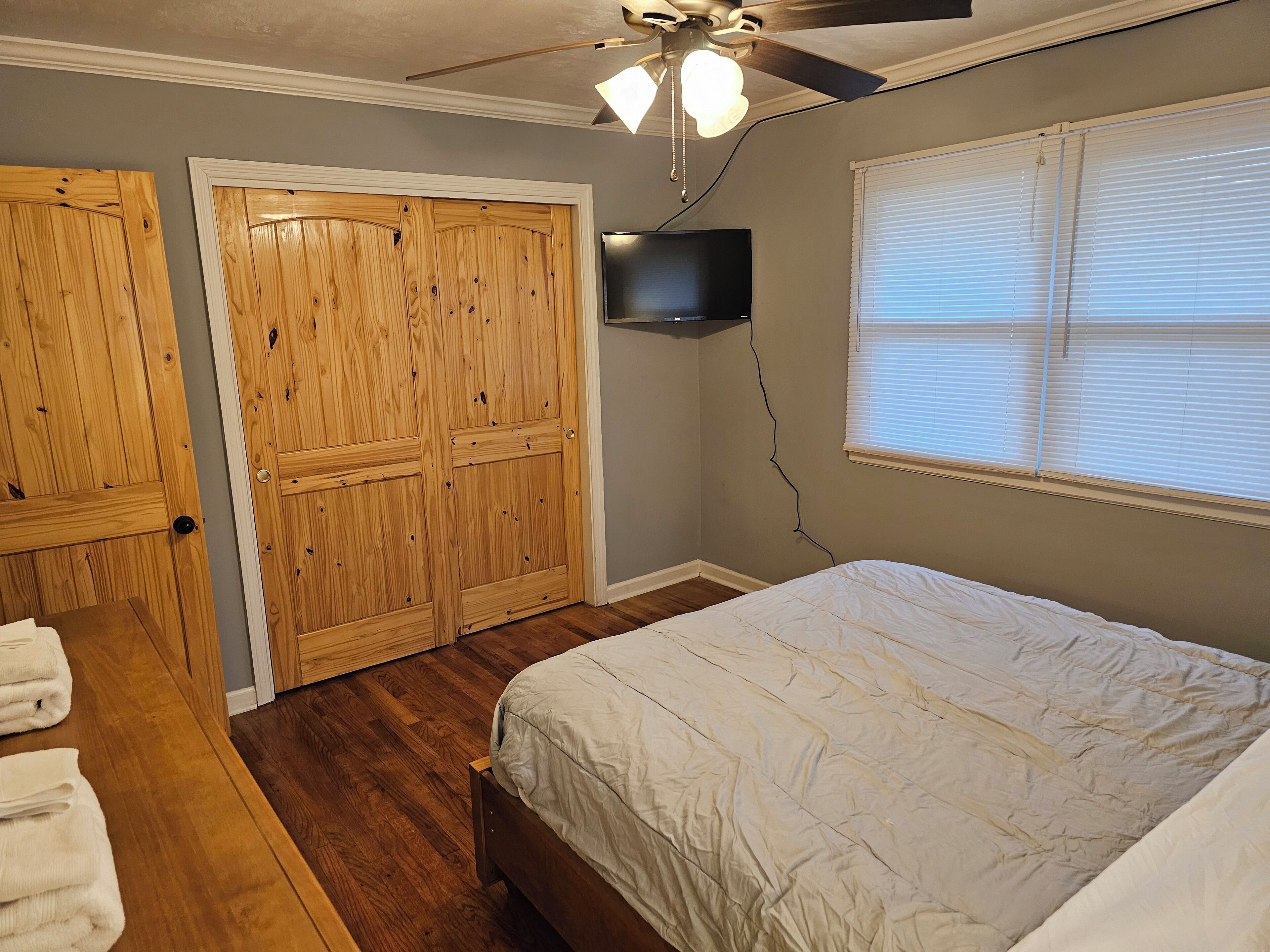 Ein Schlafzimmer mit Bett, Holzschrank, Deckenventilator, Fernseher und einem Fenster mit Jalousien.
