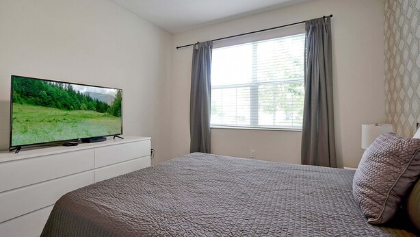 Un dormitorio con cama, televisor sobre una cómoda, ventana con cortinas y un diseño estampado en la pared.