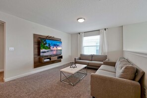 A living room with a flat-screen TV mounted on the wall, a beige sofa, a coffee table, and a window with curtains. - Family & Dog-Friendly ChampionsGate Villa | Private Pool, Game Room & Themed Rooms (Champions Gate)