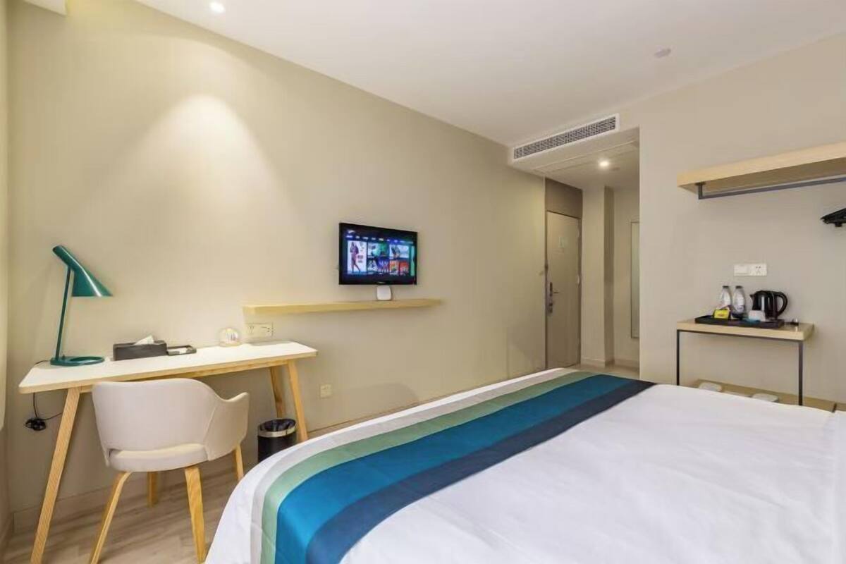 A hotel room with a bed, a desk, a chair, a TV mounted on the wall, and a small table with a kettle and cups.