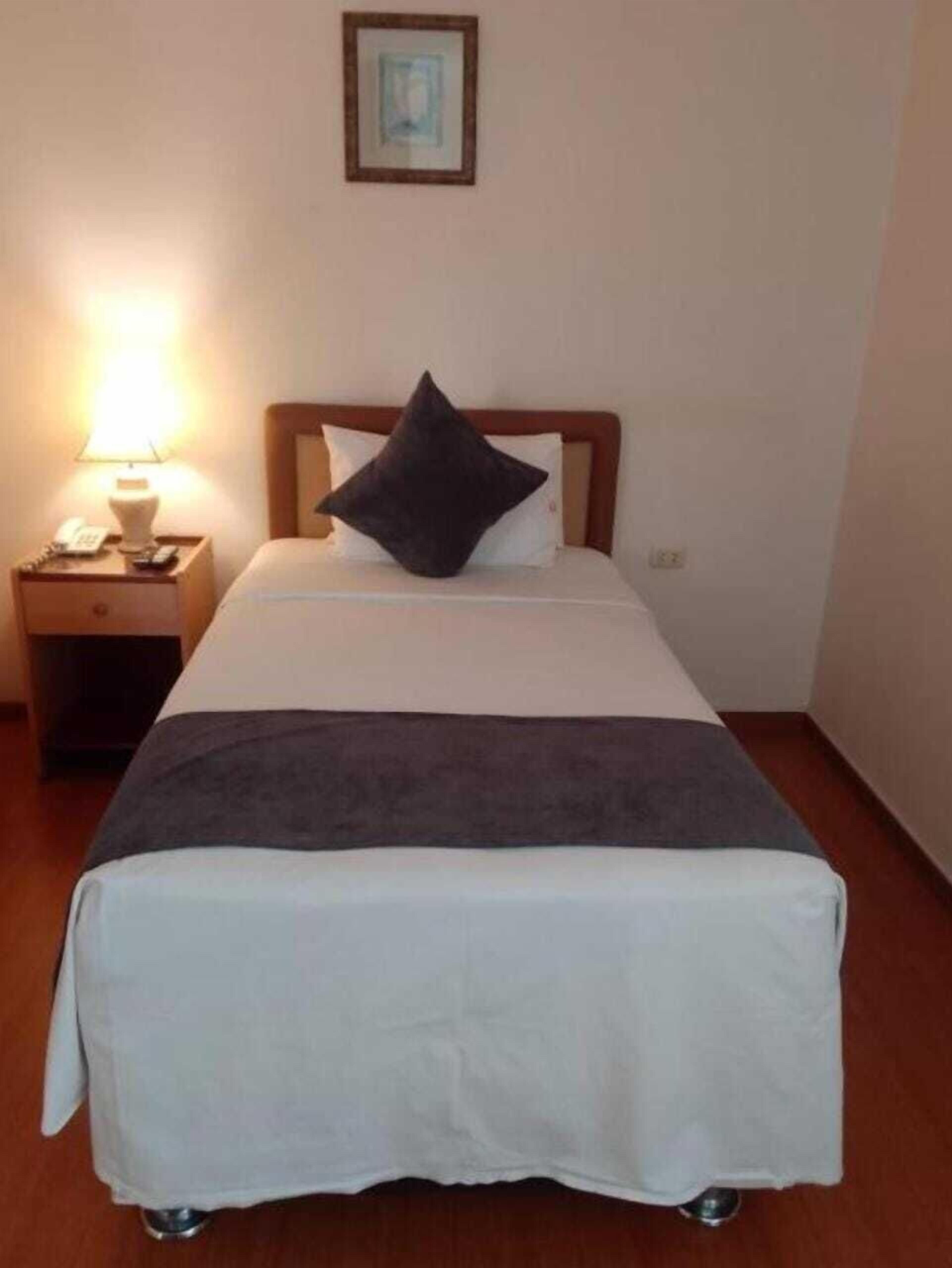 A neatly made bed with a white and gray bedspread, a dark pillow, and a wooden headboard. There is a nightstand with a lamp and a phone beside the bed.