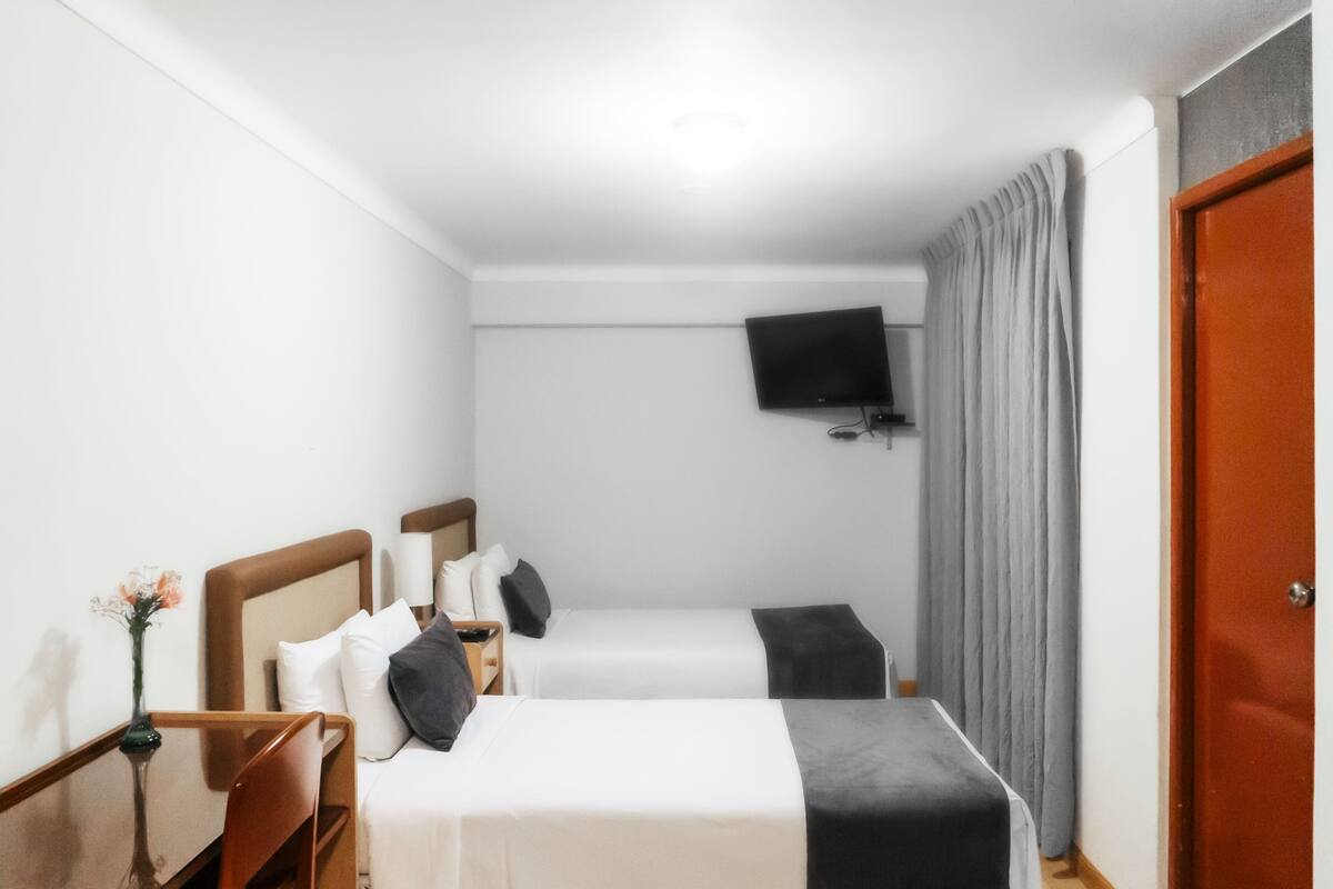 A hotel room with two single beds, a flat-screen TV mounted on the wall, a wooden nightstand, a small table with a vase, and a red door.