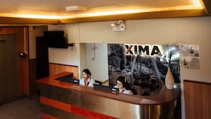 A reception desk with a 'XIMA' sign, two receptionists, and a wall-mounted television. - Xima Central Tacna (Tacna)