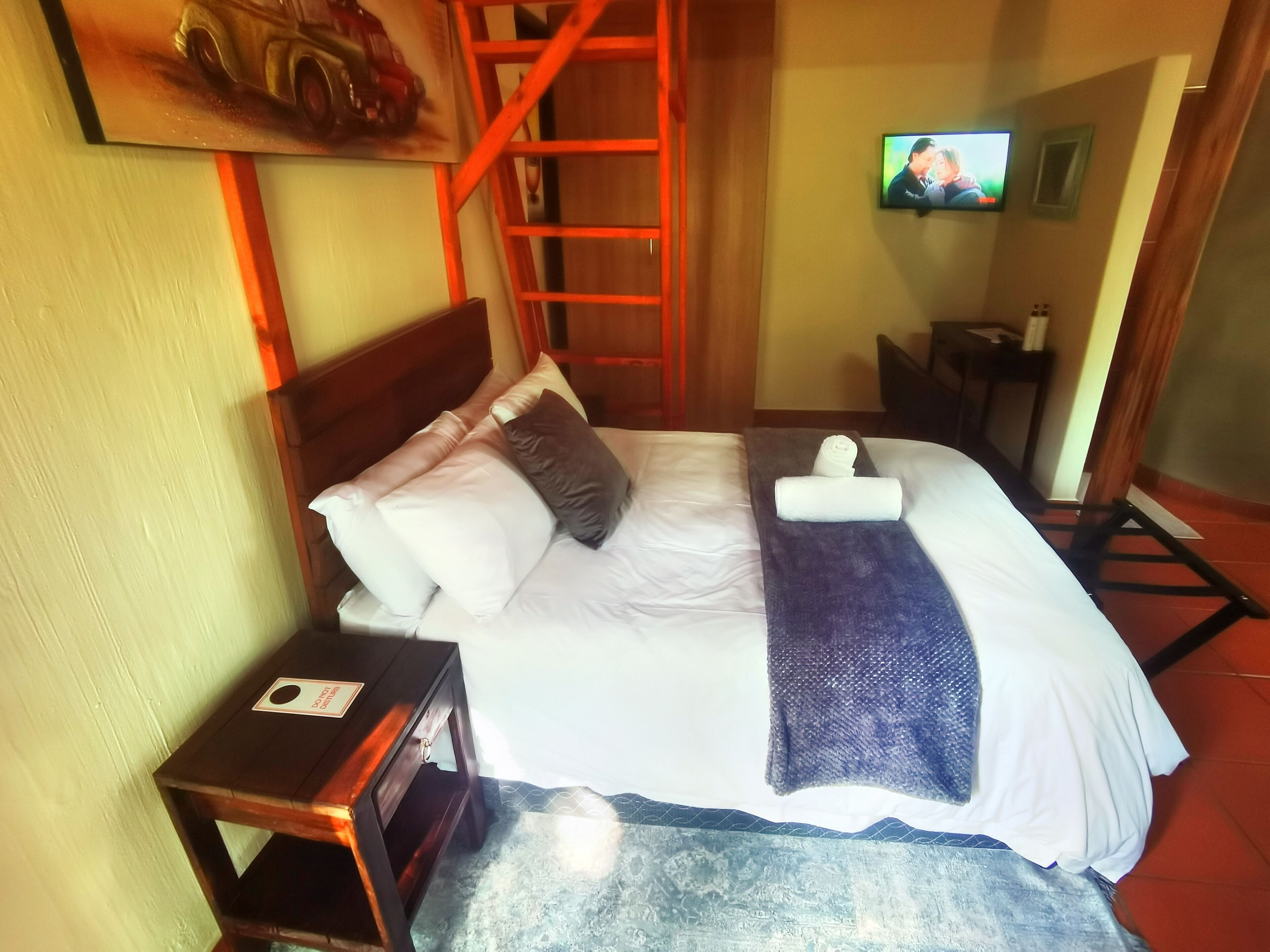 A neatly made bed with pillows and a blanket, a wooden nightstand with a card, a TV mounted on the wall, and a red ladder against the wall.