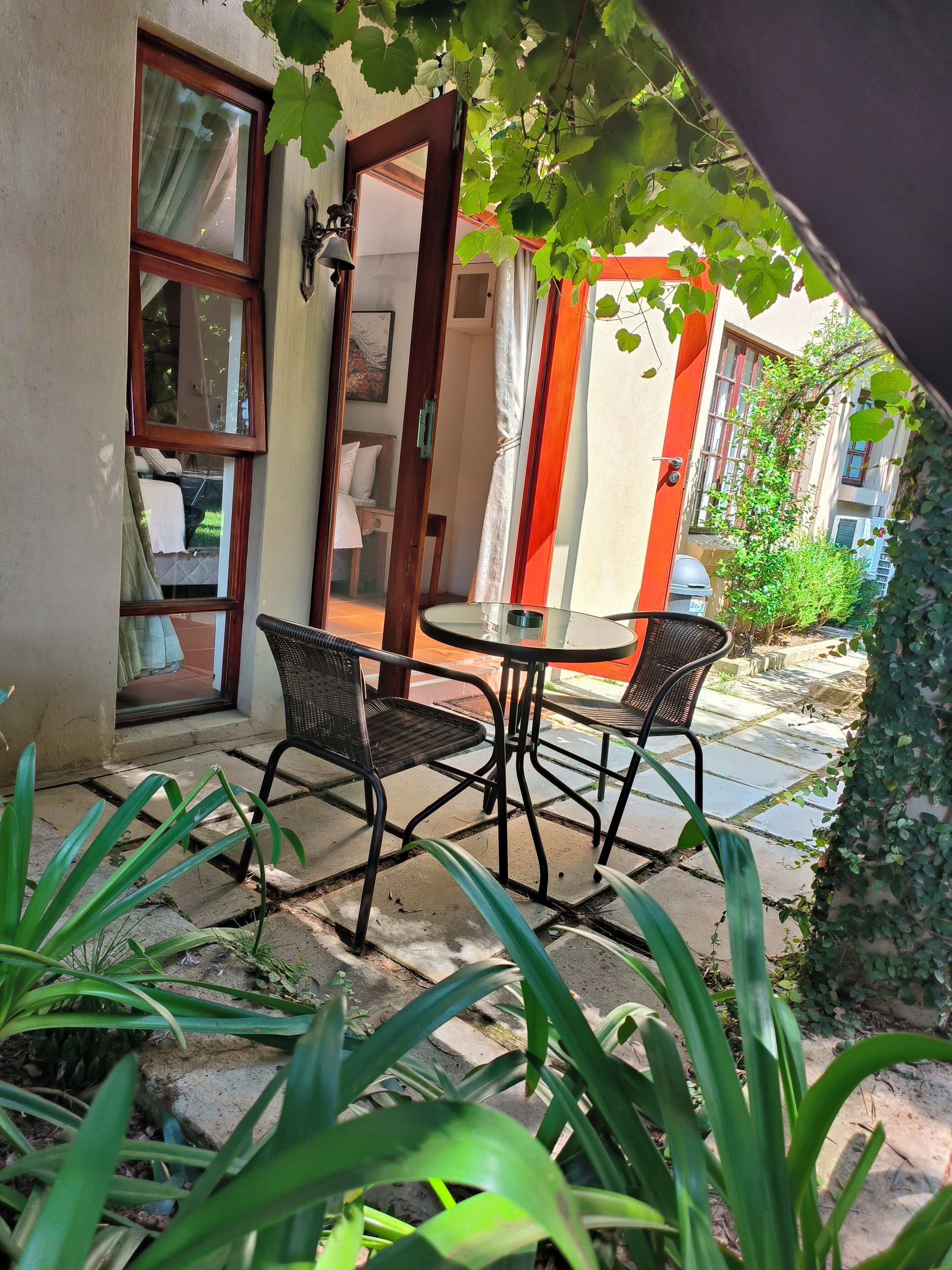 A patio with a table and chairs, surrounded by greenery and a building with a red door.