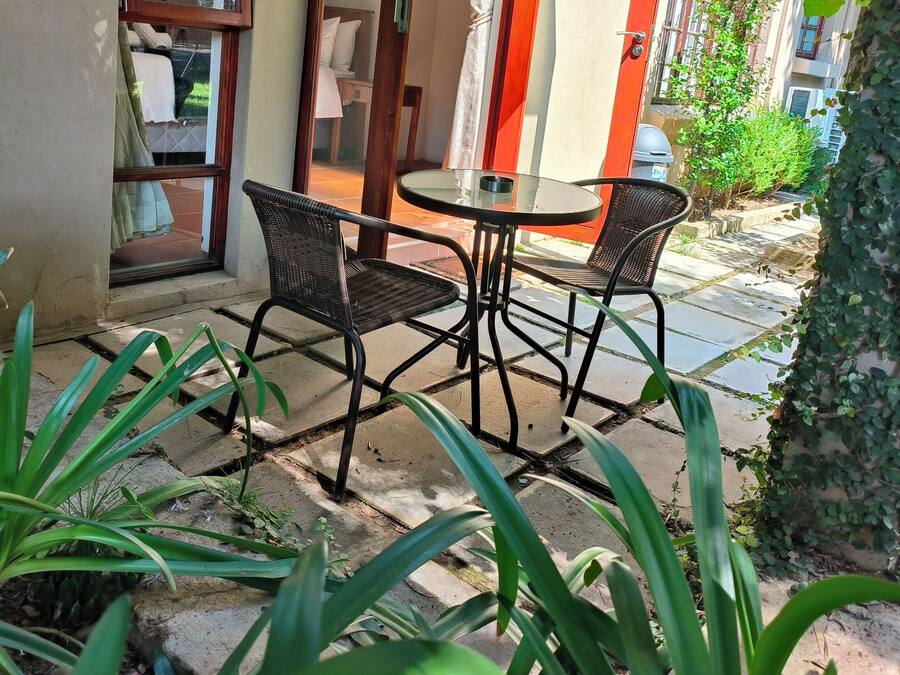 A patio with a table and chairs, surrounded by greenery and a building with a red door.