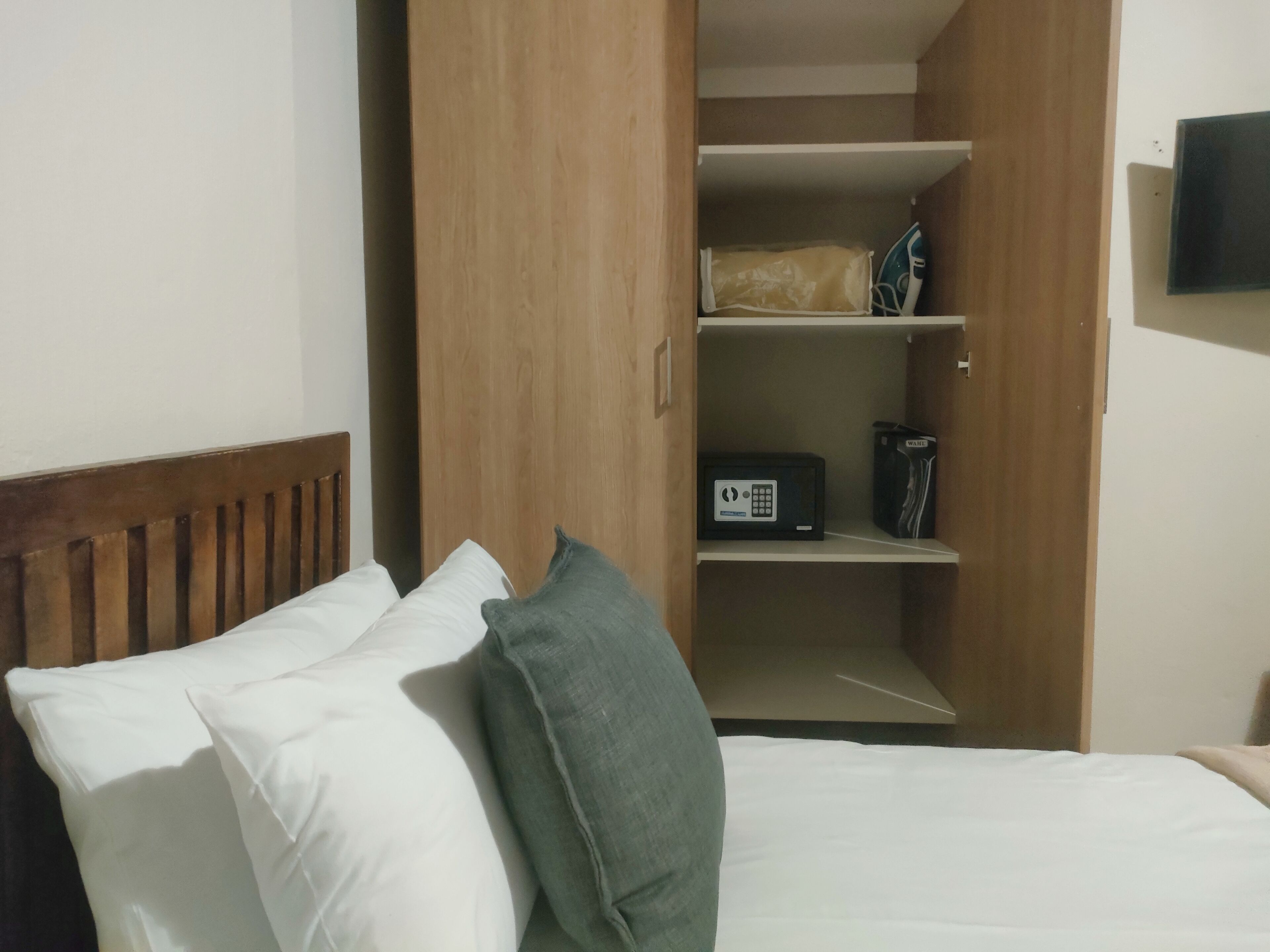 A hotel room with a bed, pillows, a wooden headboard, a wardrobe, and a television on the wall.