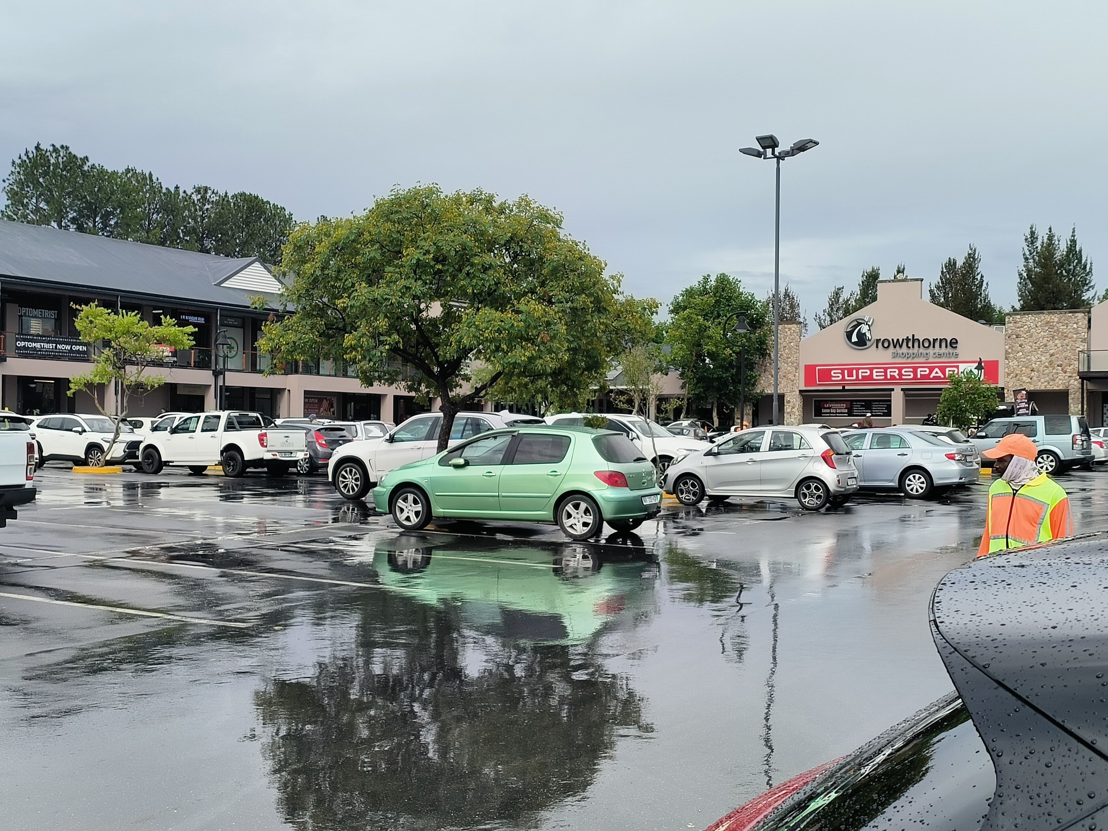 Ein Parkplatz mit verschiedenen Autos, einer Reihe Geschäfte und feuchtem Boden, der die Umgebung spiegelt.