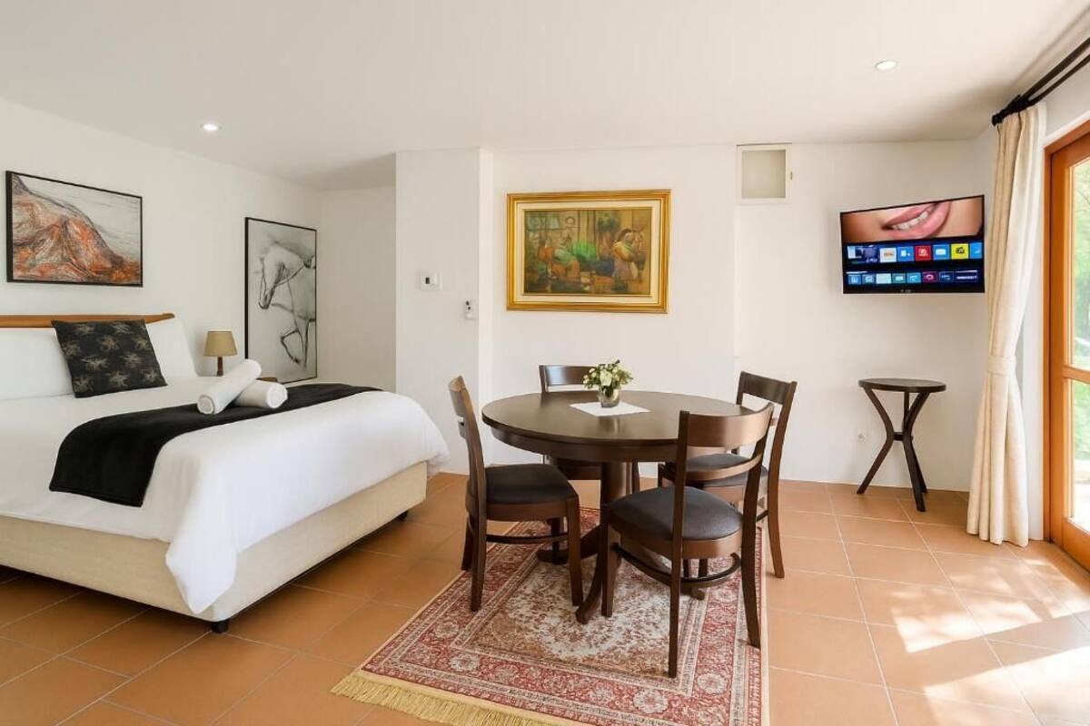 Una habitación de hotel con una cama, una mesa de comedor con sillas, un televisor montado en la pared y obras de arte en las paredes.