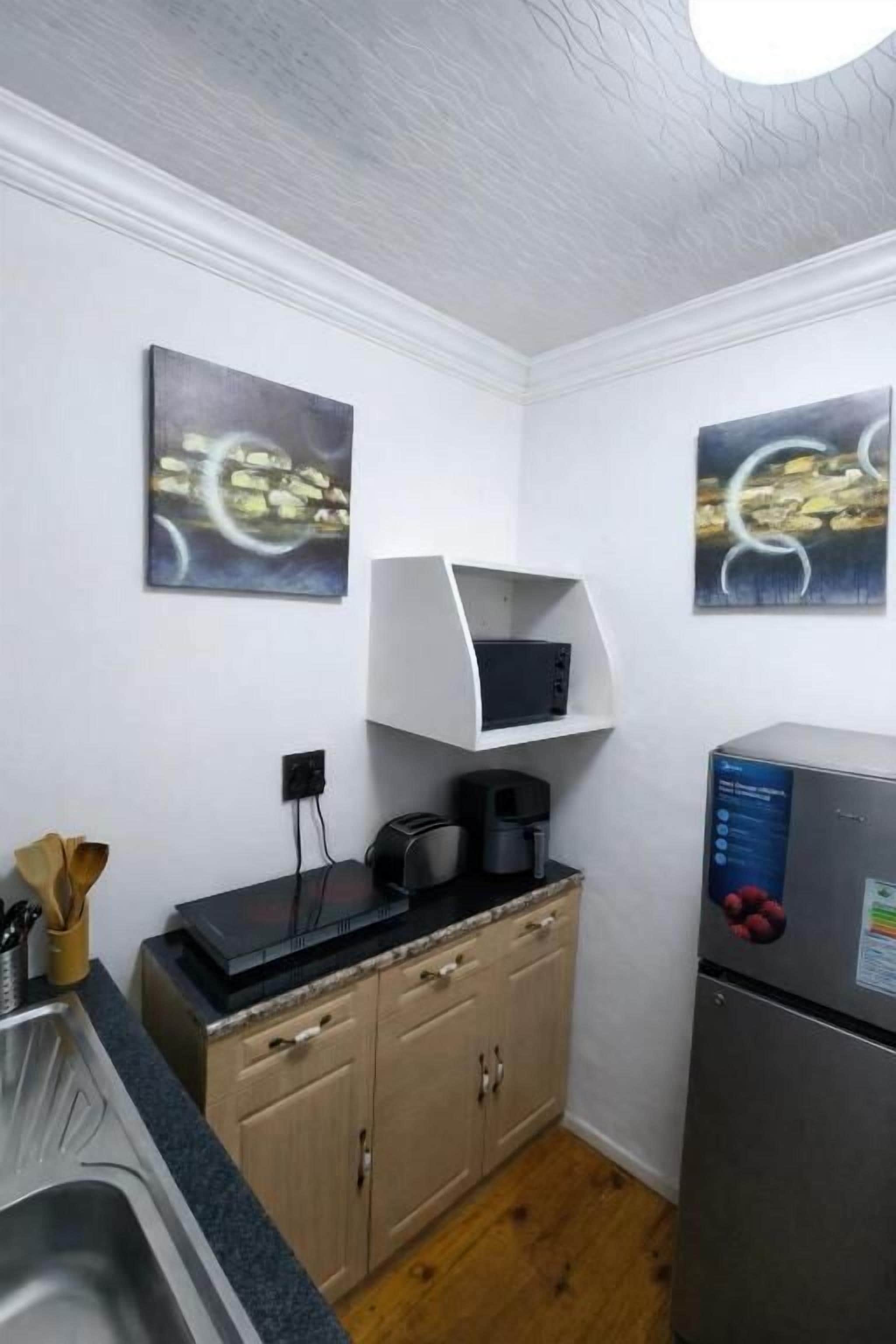 Une cuisine compacte équipée d’un four à micro-ondes, d’une machine à café et d’un réfrigérateur. Deux tableaux abstraits ornent les murs.