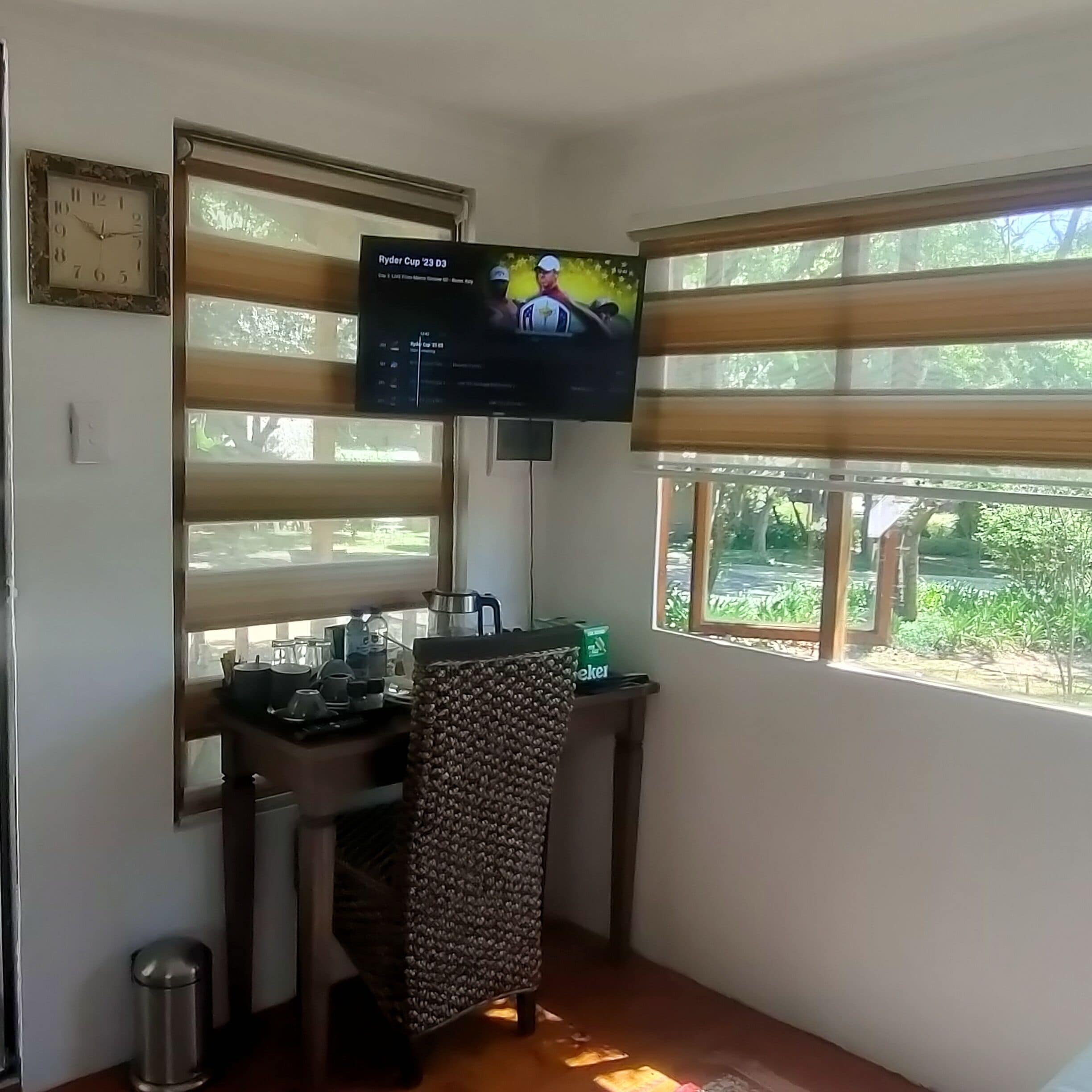 Een kleine kamer met een bureau, een aan de muur bevestigde televisie, een raam met zonwering en uitzicht op groen.
