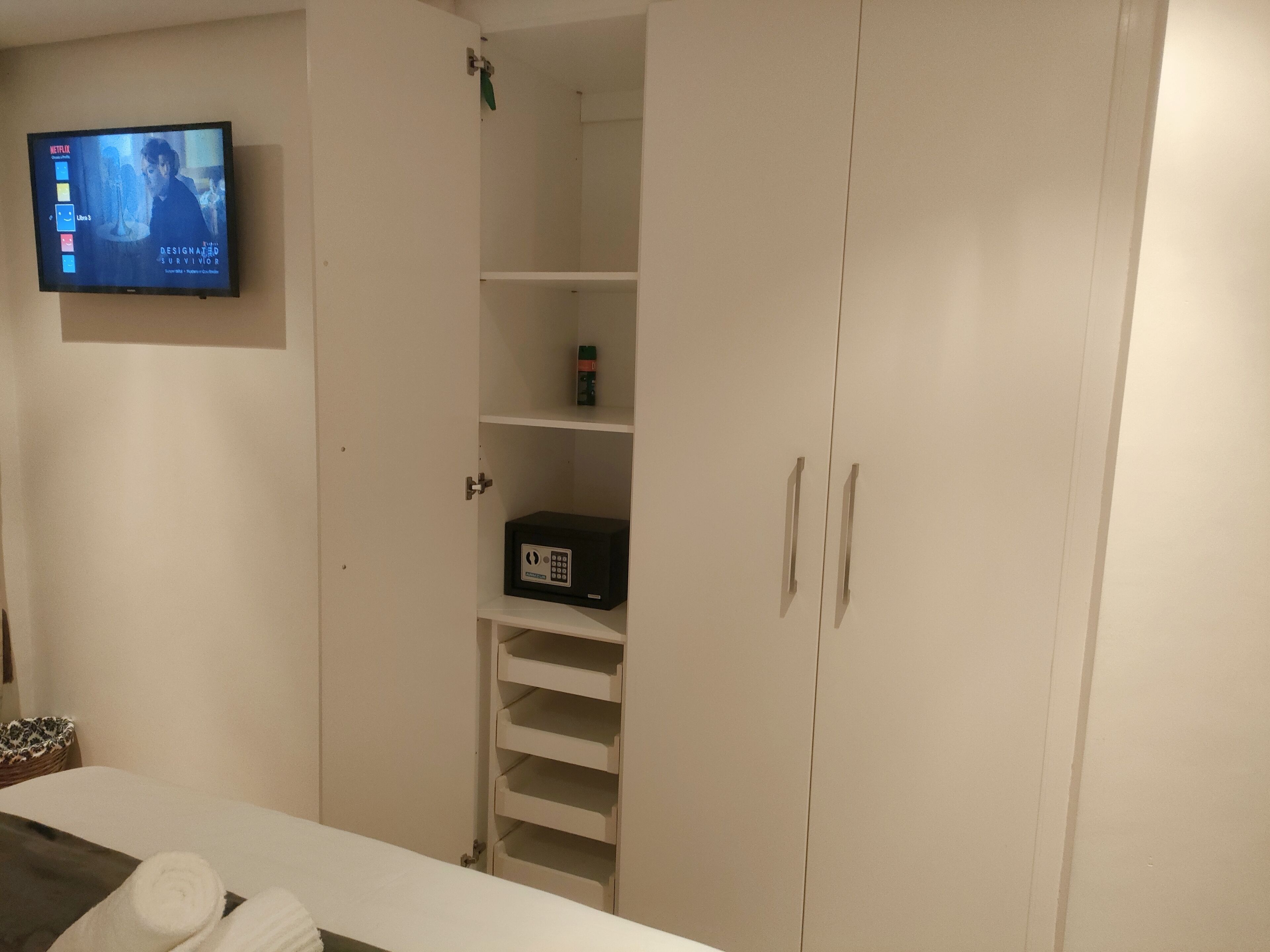 A hotel room with a white wardrobe, a television mounted on the wall, and a small safe on the lower shelf.