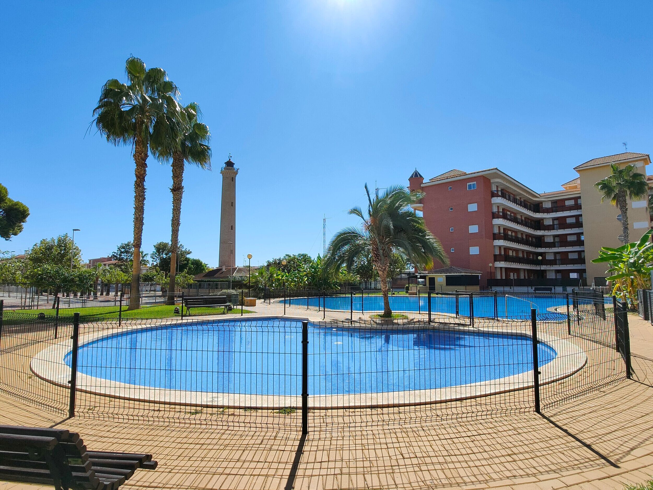 Une piscine entourée d’une clôture, avec des palmiers et une haute tour en arrière-plan.