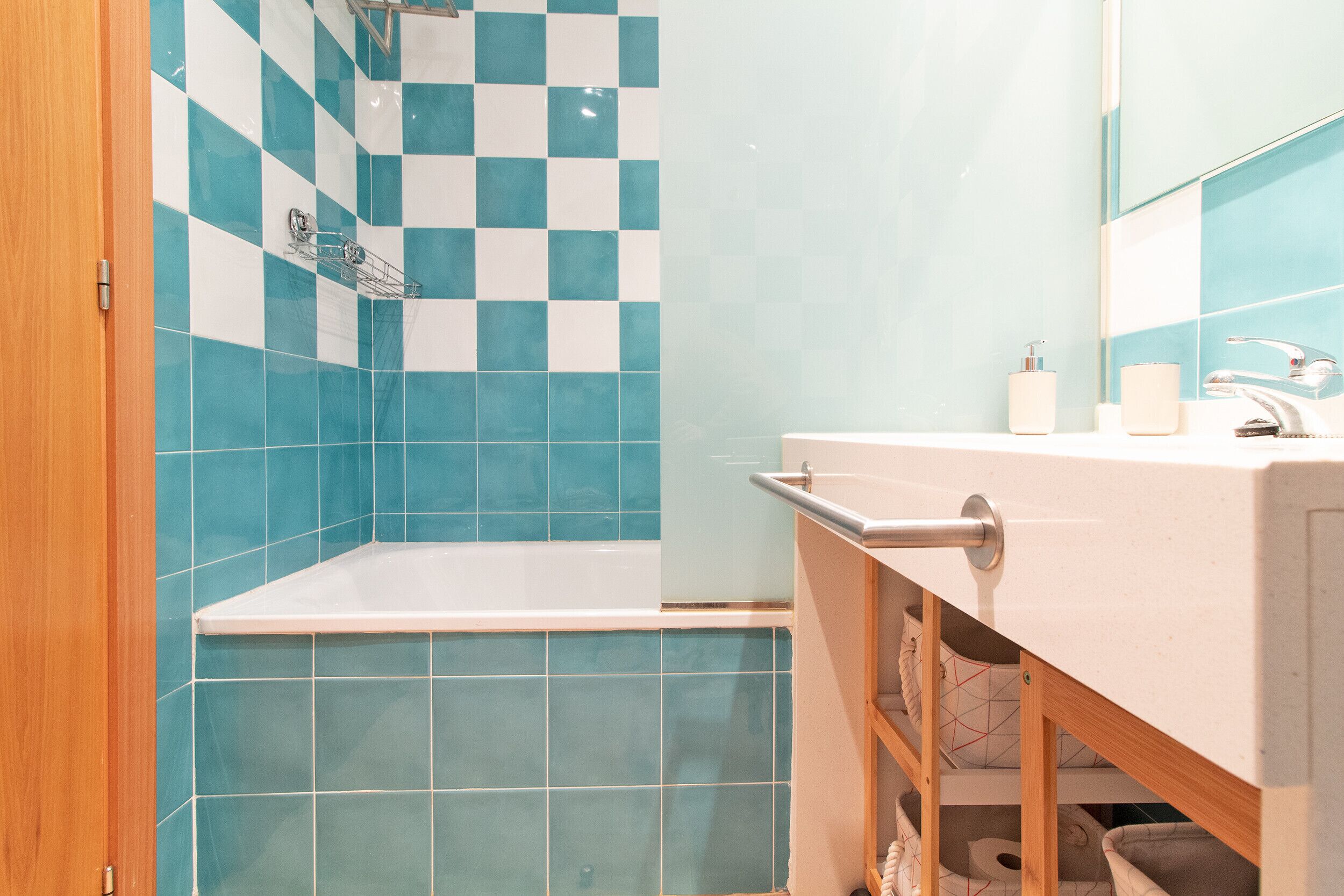 Un baño con pared de azulejos a cuadros, una bañera, un lavabo con espejo y un mueble de madera debajo.