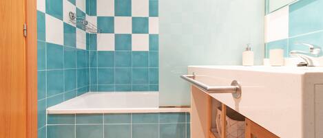 Un baño con pared de azulejos a cuadros, una bañera, un lavabo con espejo y un mueble de madera debajo.