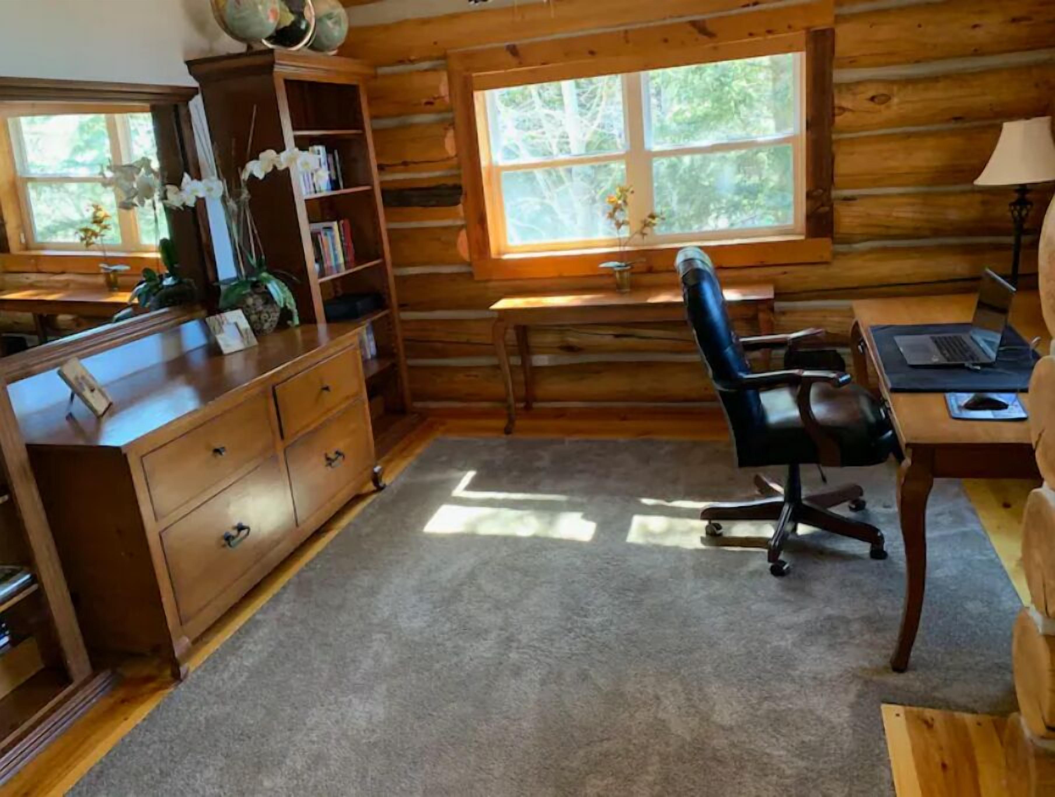 L’intérieur d’une cabane en rondins, avec un bureau en bois, une chaise, une bibliothèque et une fenêtre donnant sur la forêt.