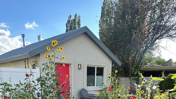 Un camino pavimentado que lleva a una casa con puerta roja, rodeada de diversas plantas en flor y una bicicleta.