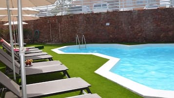 A swimming pool with lounge chairs and umbrellas, surrounded by a stone wall and green lawn.