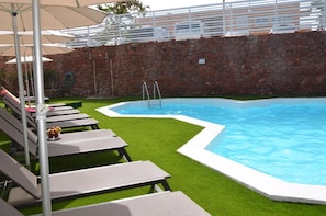 Piscina com espreguiçadeiras e guarda-sóis, cercada por um muro de pedra e gramado verde.