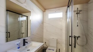 Une salle de bain avec un lavabo blanc, un miroir, des toilettes et une douche dotée d’une porte en verre, ainsi qu’une fenêtre.