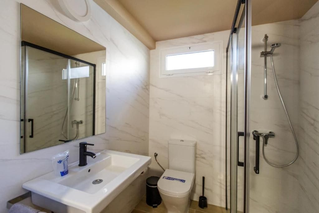 A bathroom with a white sink, a mirror, a toilet, a shower with a glass door, and a window.